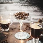 Gorgeous coffee glasses filled with hot coffee and cocktail glass filled with coffee beans standing on dark ground, surrounded by coffee beans.