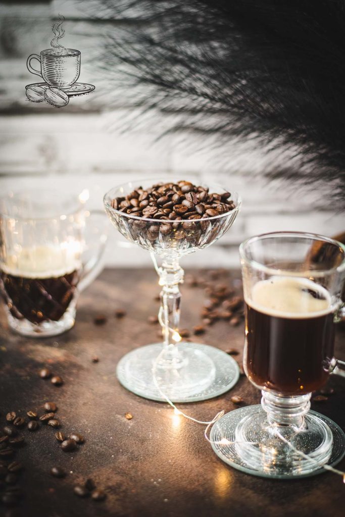 Gorgeous coffee glasses filled with hot coffee and cocktail glass filled with coffee beans standing on dark ground, surrounded by coffee beans.