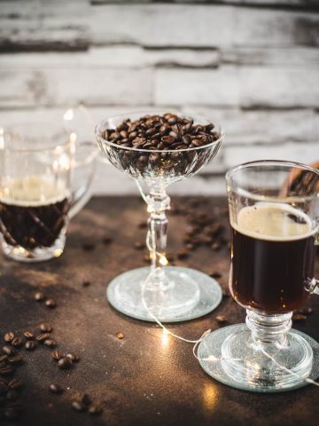 Gorgeous coffee glasses filled with hot coffee and cocktail glass filled with coffee beans standing on dark ground, surrounded by coffee beans.