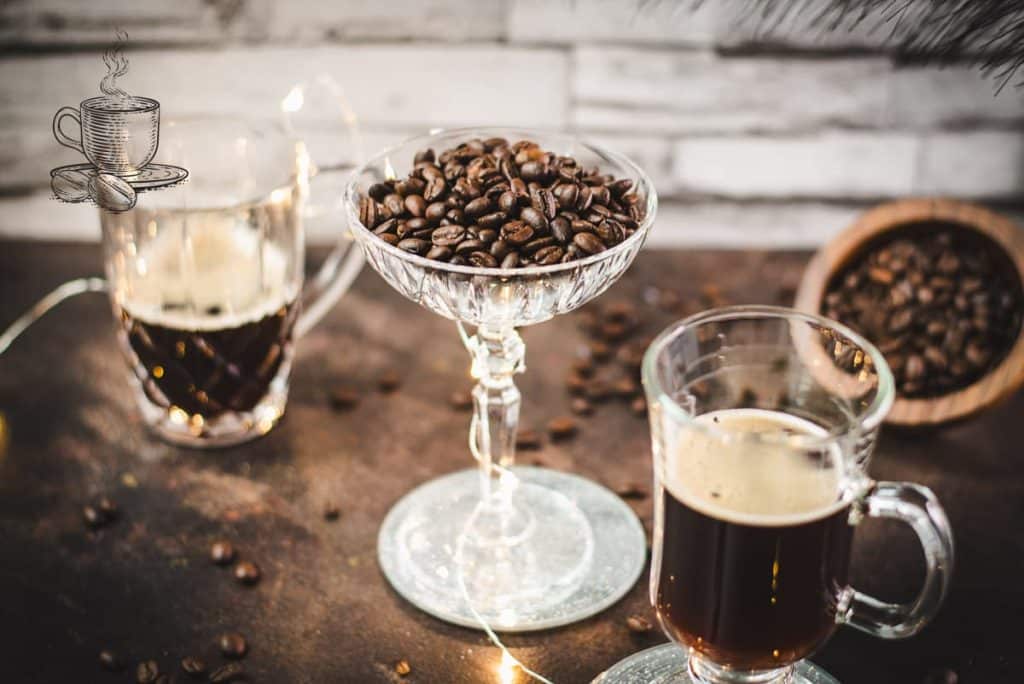 Gorgeous coffee glasses filled with hot coffee and cocktail glass filled with coffee beans standing on dark ground, surrounded by coffee beans.