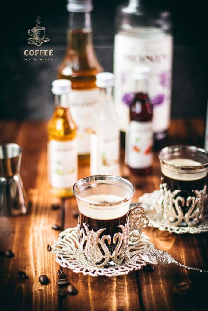 Two cups of coffee placed in front of a variety of delicious syrup bottles.