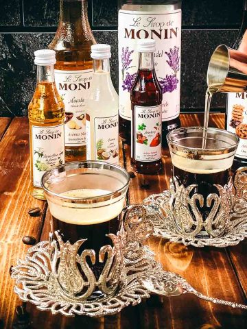 Two cups of coffee placed in front of a variety of delicious syrup bottles. Syrup is poured into coffee glass using a jigger.