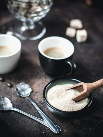 Brown sugar in dark green bowl next to black cup filled with black coffee.