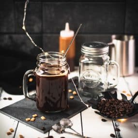 Mason jar filled with delicious coffee caramel. Placed on black slate, surrounded by coffee beans and caramel pieces.