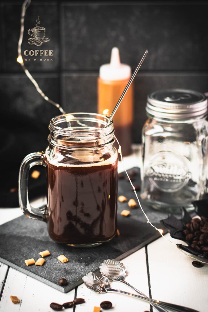 Mason jar filled with delicious coffee caramel. Placed on black slate, surrounded by coffee beans and caramel pieces.