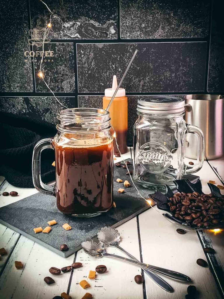 Mason jar filled with delicious coffee caramel. Placed on black slate, surrounded by coffee beans and caramel pieces.