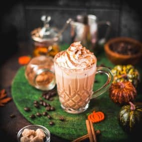 Coffee glass filled with delicious pumpkin spiced latte, garnished with whipped cream and dusted with a cocoa cinnamon mix. Placed on green felt.