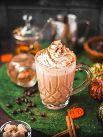 Coffee glass filled with delicious pumpkin spiced latte, garnished with whipped cream and dusted with a cocoa cinnamon mix. Placed on green felt.