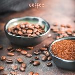 Two bowls each filled with coffee beans and grated coffee on rust brown background surrounded by coffee beans.