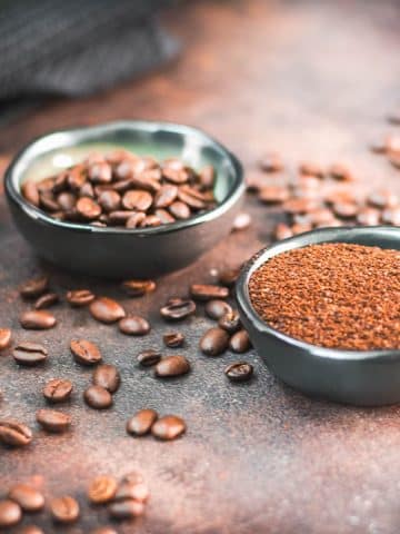 Two bowls each filled with coffee beans and grated coffee on rust brown background surrounded by coffee beans.