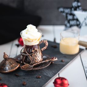 Gorgeous mediaeval coffee glass filled with black coffee, topped with eggnog flavored whipped cream. Placed on black slate plate next to a glass of eggnog.