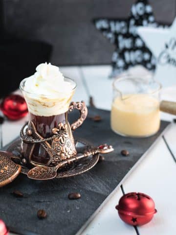 Gorgeous mediaeval coffee glass filled with black coffee, topped with eggnog flavored whipped cream. Placed on black slate plate next to a glass of eggnog.