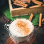 Gorgous cinnamon dolce latte, dusted with ground cinnamon. Placed on wooden coaster with cinnamon sticks in the background.