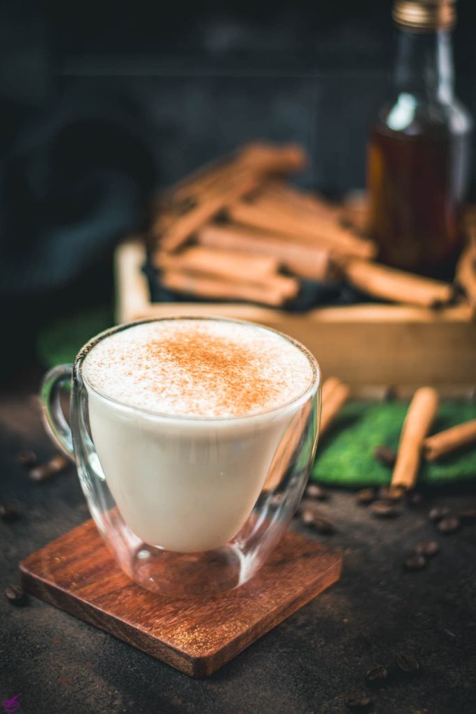 Gorgous cinnamon dolce latte, dusted with ground cinnamon. Placed on wooden coaster with cinnamon sticks in the background.