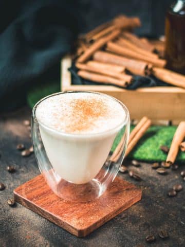 Gorgous cinnamon dolce latte, dusted with ground cinnamon. Placed on wooden coaster with cinnamon sticks in the background.