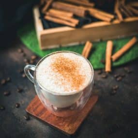 Gorgous cinnamon dolce latte, dusted with ground cinnamon. Placed on wooden coaster with cinnamon sticks in the background.