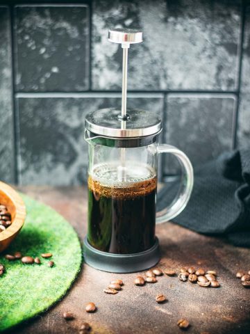 French press filled with cold water and ground coffee next to a bowl of coffee beans - an easy way to make cold brew coffee!