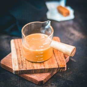 Glass container filled with delicious homemade caramel sauce placed on wooden coaster.