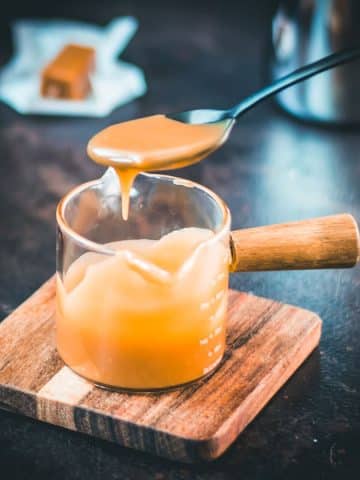 Spoon with homemade caramel sauce hovering over glass jar filled with caramel sauce.