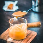 Spoon with homemade caramel sauce hovering over glass jar filled with caramel sauce.