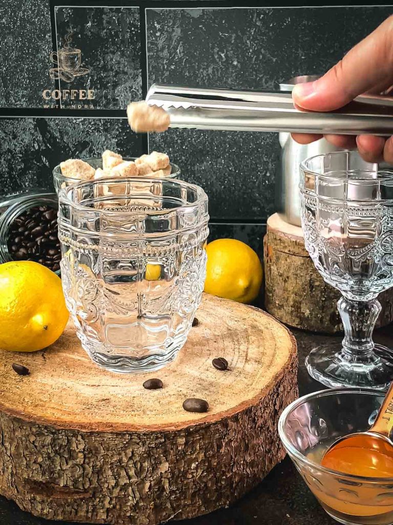 A piece of brown sugar is placed in a beautiful glass using tongs.