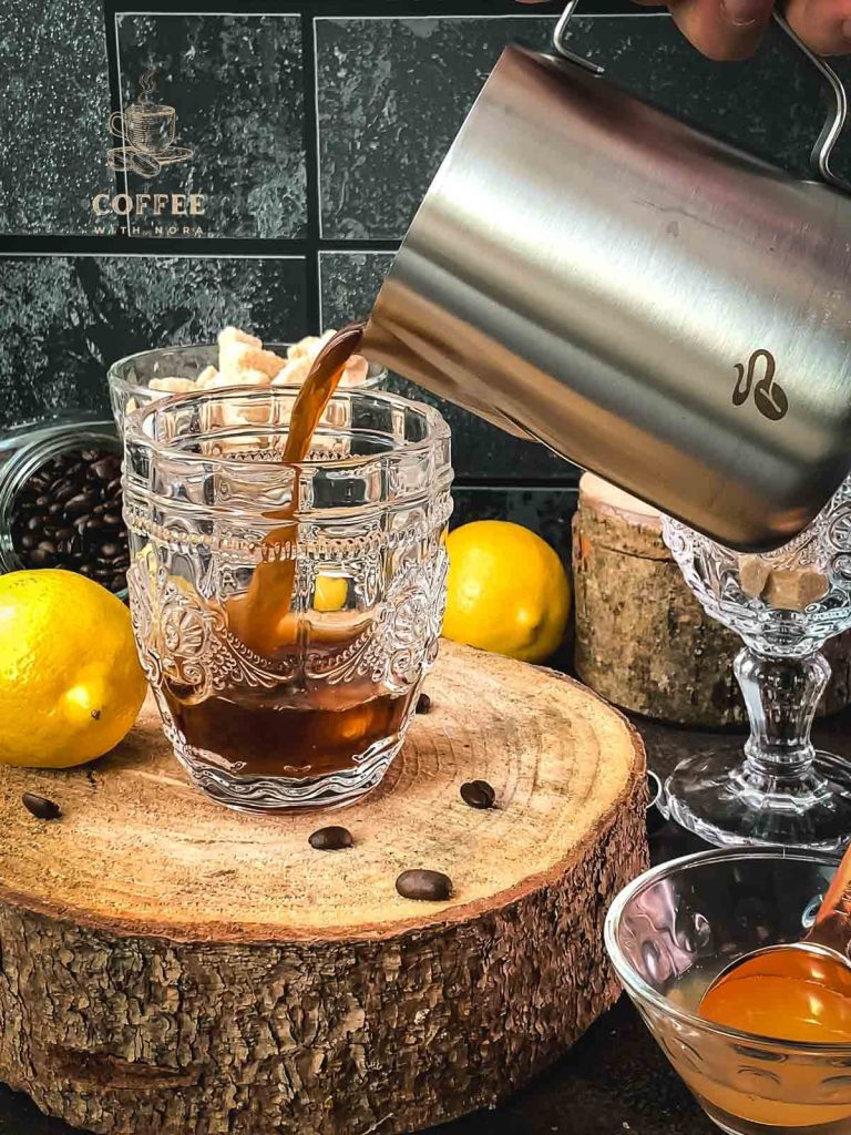 Coffee is poured into a serving glass using a metal pitcher.