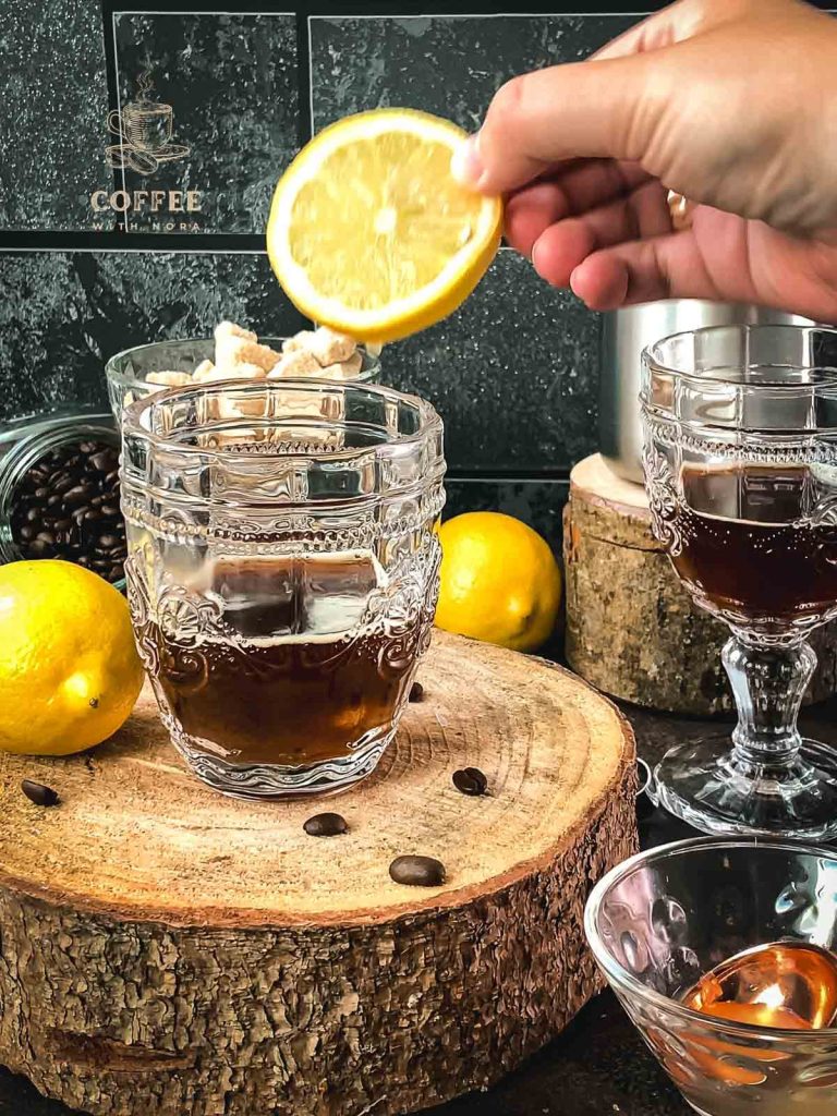 One hand holds the slice of lemon and places it in a glass of coffee.