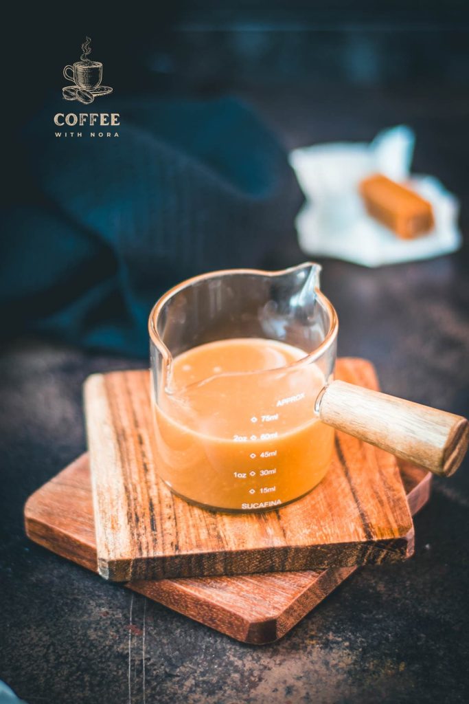 Glass container filled with delicious homemade caramel sauce placed on wooden coaster.