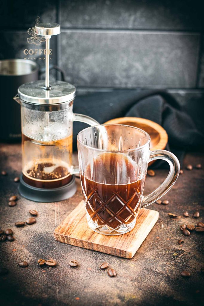 Gorgous glass filled with French press coffee placed on wooden coaster. In the back an empty French press coffee maker.