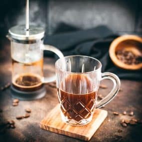 Gorgous glass filled with French press coffee placed on wooden coaster. In the back an empty French press coffee maker.