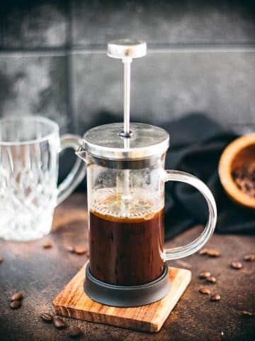 French press coffee maker filled with coffee on a wooden coaster. In the back an empty coffee glass.