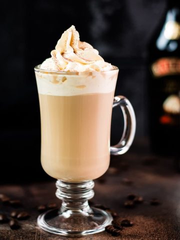 Gorgeous glass filled with scrumptious Bailey's coffee, topped with whipped cream and dusted with cocoa. Placed on dark ground in front of an Salted Baileys Caramel bottle.