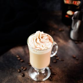 Gorgeous glass filled with scrumptious Bailey's coffee, topped with whipped cream and dusted with cocoa. Placed on dark ground in front of an Salted Baileys Caramel bottle.