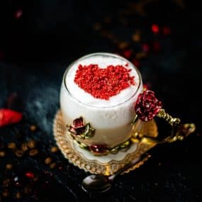 Beautiful coffee glass decorated with a red and gold butterfly and flowers. Filled with delicious strawberry latte and topped with heart-shaped freeze-dried strawberry powder. Placed on dark ground.