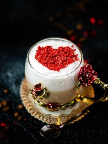 Beautiful coffee glass decorated with a red and gold butterfly and flowers. Filled with delicious strawberry latte and topped with heart-shaped freeze-dried strawberry powder. Placed on dark ground.