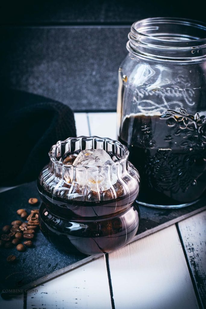 Gorgeous glass filled with cubed ice and cold brew coffee. Placed on a black slate plate on white wooden ground.