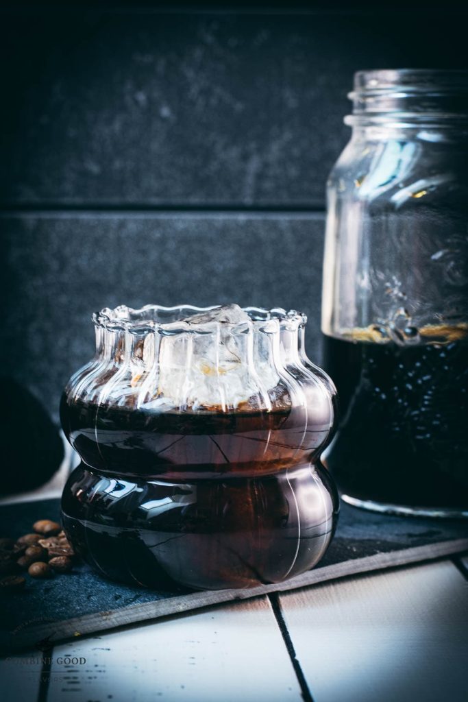 Gorgeous glass filled with cubed ice and cold brew coffee. Placed on a black slate plate on white wooden ground.