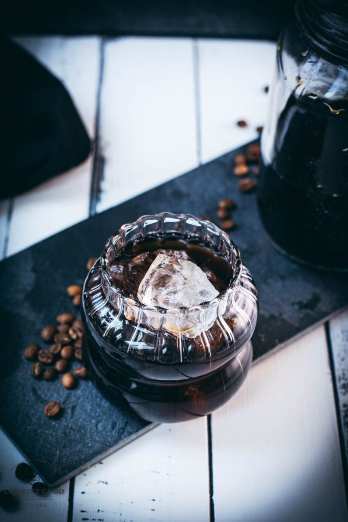 Gorgeous glass filled with cubed ice and cold brew coffee. Placed on a black slate plate on white wooden ground.