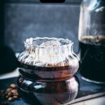 Gorgeous glass filled with cubed ice and cold brew coffee. Placed on a black slate plate on white wooden ground.