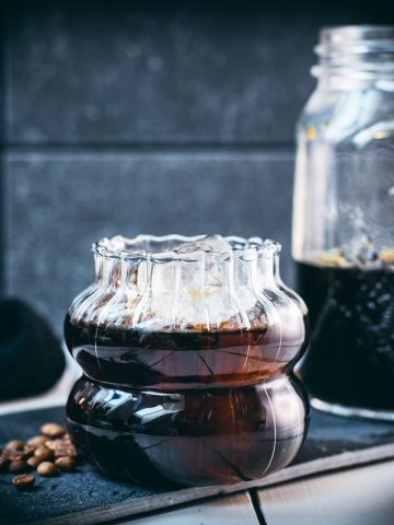 Gorgeous glass filled with cubed ice and cold brew coffee. Placed on a black slate plate on white wooden ground.