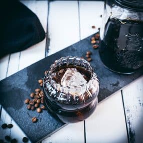 Gorgeous glass filled with cubed ice and cold brew coffee. Placed on a black slate plate on white wooden ground.