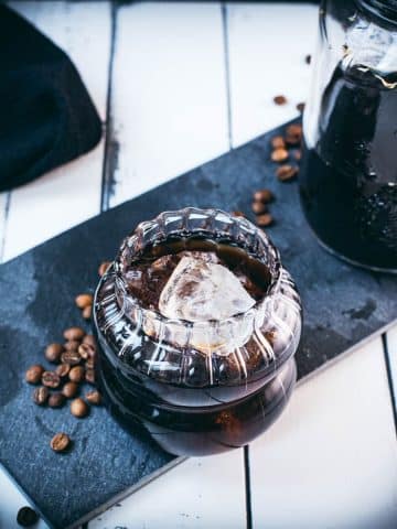 Gorgeous glass filled with cubed ice and cold brew coffee. Placed on a black slate plate on white wooden ground.