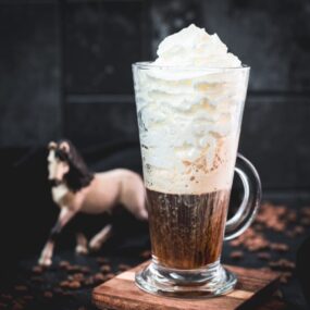 Beautiful tall glass filled with strong coffee and topped with whipped cream. Placed on brown coasters and pretty horse in the background. The Einspänner, a Viennese classic!