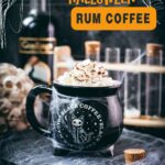 Spooky cauldron coffee mug filled with delicious coffee with rum. Topped with sweetened whipped cream and placed on a black plate surrounded by cobweb.