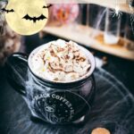 Spooky cauldron coffee mug filled with delicious coffee with rum. Topped with sweetened whipped cream and placed on a black plate surrounded by cobweb.