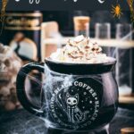 Spooky cauldron coffee mug filled with delicious coffee with rum. Topped with sweetened whipped cream and placed on a black plate surrounded by cobweb.