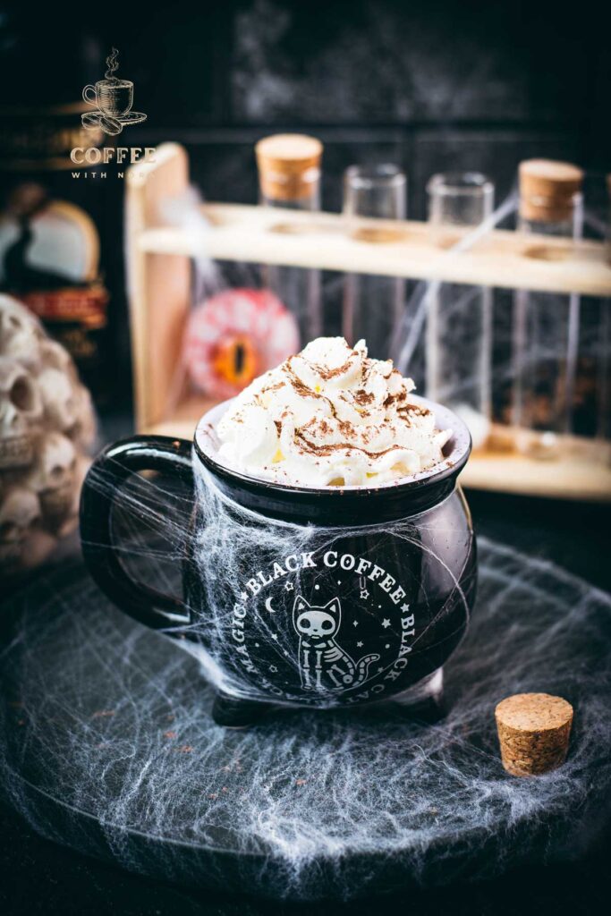 Spooky cauldron coffee mug filled with delicious coffee with rum. Topped with sweetened whipped cream and placed on a black plate surrounded by cobweb.