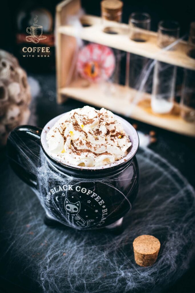 Spooky cauldron coffee mug filled with delicious coffee with rum. Topped with sweetened whipped cream and placed on a black plate surrounded by cobweb.