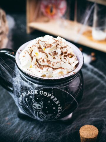 Spooky cauldron coffee mug filled with delicious coffee with rum. Topped with sweetened whipped cream and placed on a black plate surrounded by cobweb.