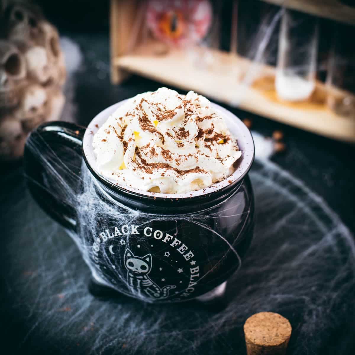 Spooky cauldron coffee mug filled with delicious coffee with rum. Topped with sweetened whipped cream and placed on a black plate surrounded by cobweb.